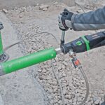 Worker uses a core drilling machine to bore through concrete with a green diamond core bit at a construction site.