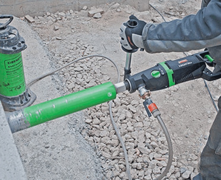 Worker uses a core drilling machine to bore through concrete with a green diamond core bit at a construction site.