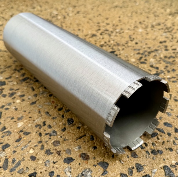 Silver diamond core drill bit with segmented cutting teeth rests on a speckled workshop surface for display.